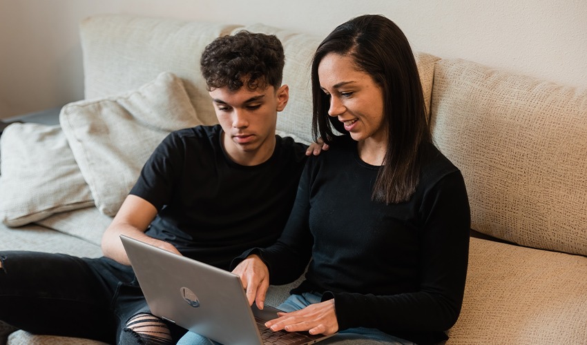 A teenager and his parent sat on a sofa looking at a laptop.