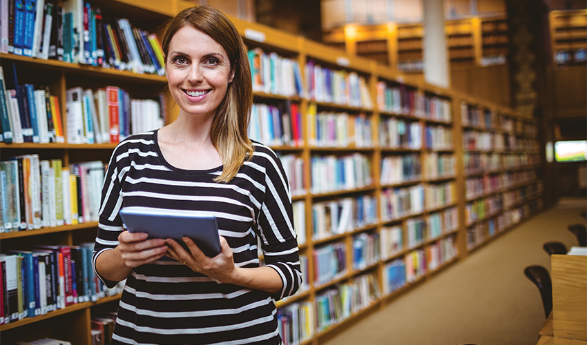 A smiling woman standing in a library holding a tablet, with bookshelves filled with books in the background.