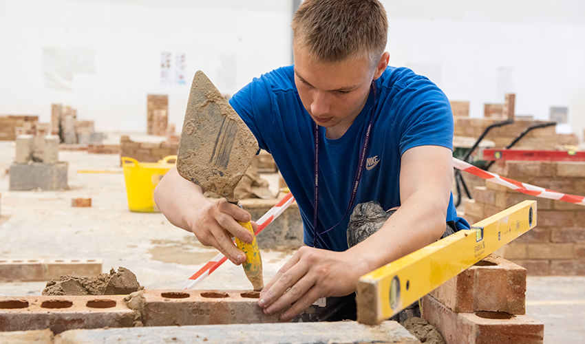 An image of a student building a brick wall, holding a trowel tapping a brick into place.
