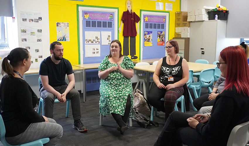 A group of adult learners practicing sign language, sat on chairs in a semi-circle around their tutor.