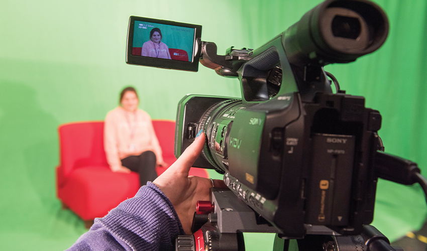 An image of a camera recording a student in a tv studio, with a big green screen behind.