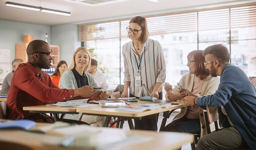 A group of adults sitting around classroom tables engaged in a collaborative activity, with an instructor standing and smiling as she interacts with them.