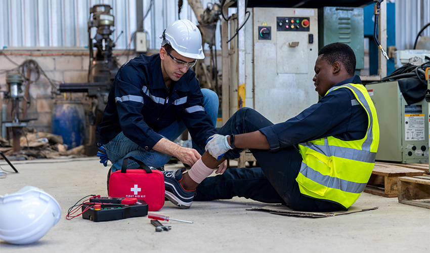 A worker wearing a hard hat wraps a bandage around a colleague’s injured ankle on the floor of an industrial workshop, with a first aid kit open beside them.