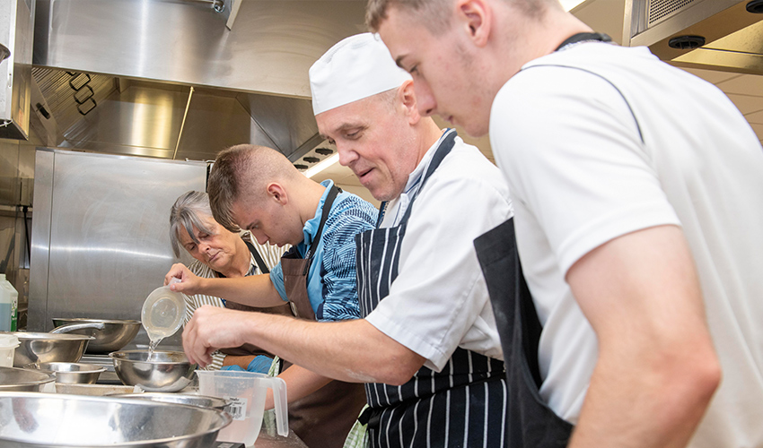 An image of chefs and students cooking and preparing ingredients in a kitchen.