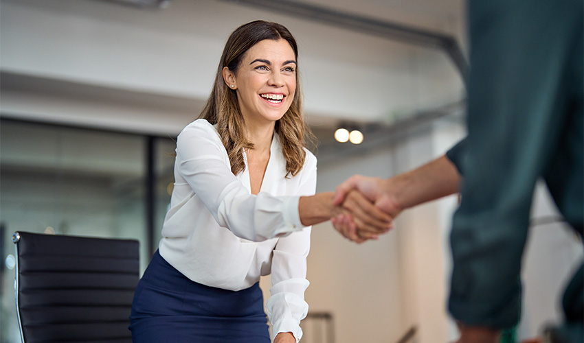 A smiling woman in business attire, in an office, leaning forward to shake hands with a man.