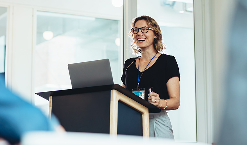 A smiling woman standing behind a podium with a laptop, giving a talk.