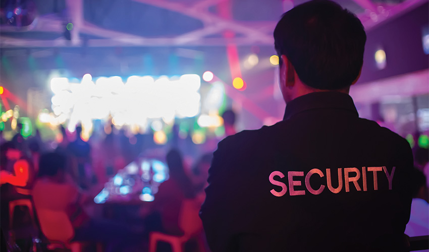 A man in a security jacket, standing in front of a club setting with lots of people and bright, colourful lights.