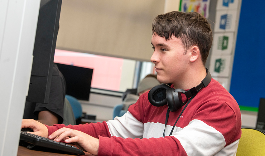 An image of a learner using a pc with headphones around his neck.