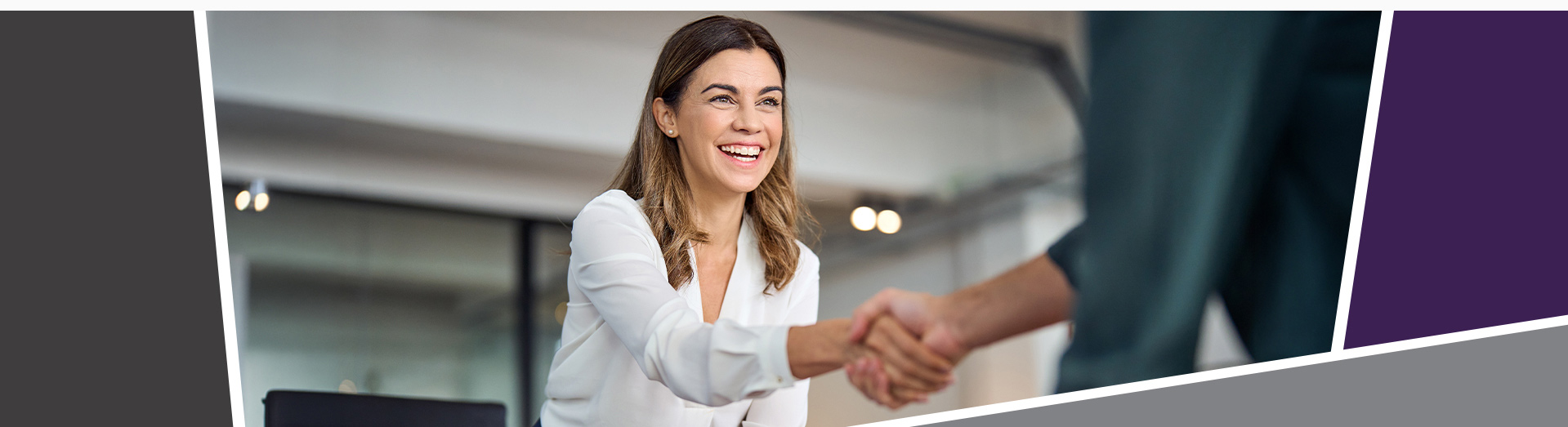 A smiling woman in business attire, in an office, leaning forward to shake hands with a man.