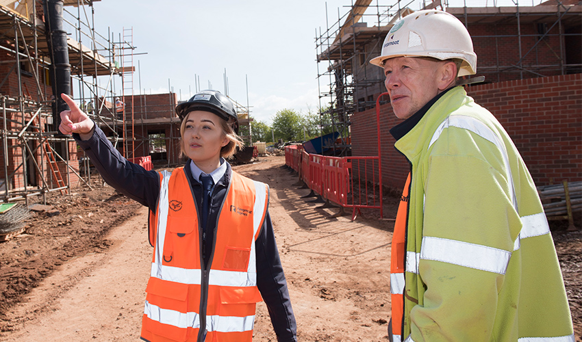 A female student in a hi-vis vest and hard hat pointing in the distance to some construction work being done on a house, with a male construction worker to her right looking in the direction she is pointing.