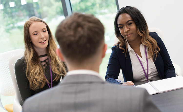 Image of two young women sat at a desk giving an interview towards a male with their back towards the camera.