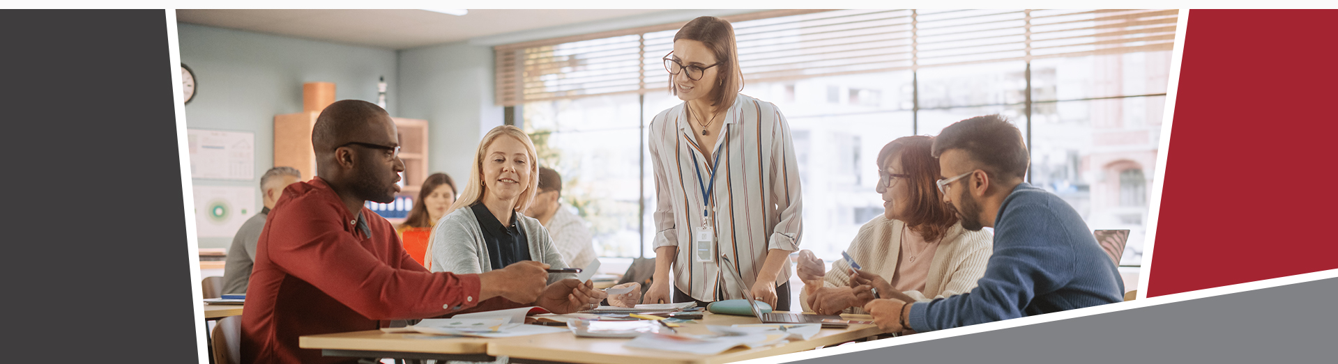A group of adults sitting around classroom tables engaged in a collaborative activity, with an instructor standing and smiling as she interacts with them.