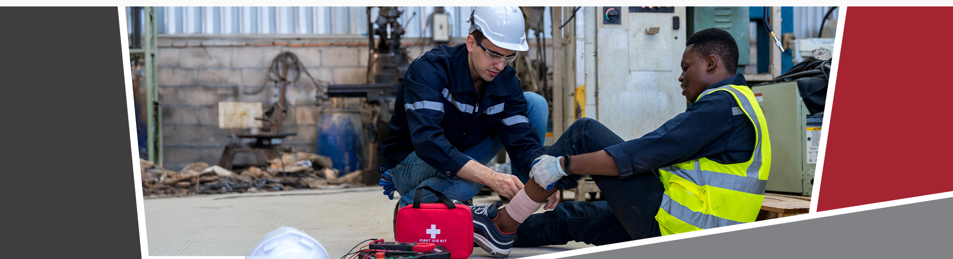 A worker wearing a hard hat wraps a bandage around a colleague’s injured ankle on the floor of an industrial workshop, with a first aid kit open beside them.