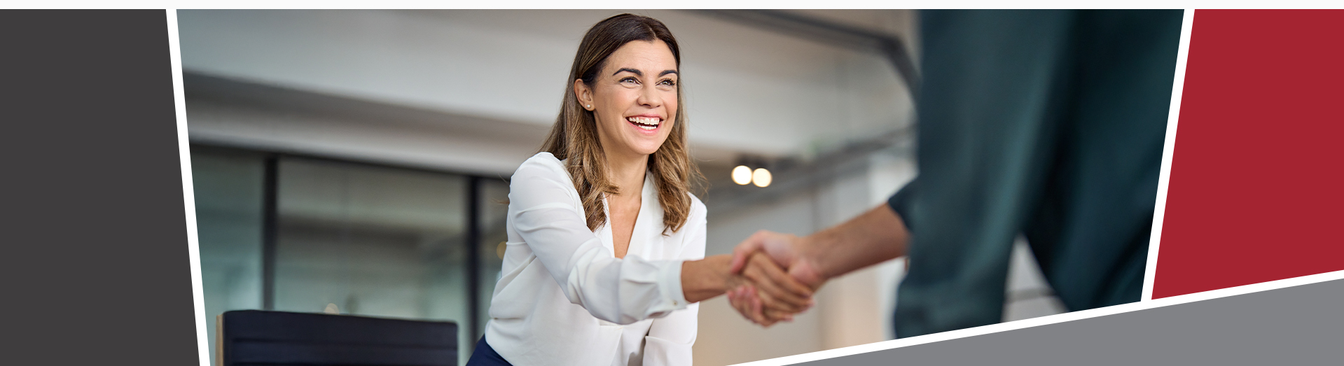 A smiling woman in business attire, in an office, leaning forward to shake hands with a man.