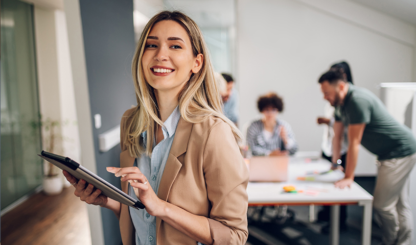 An adult woman with a tablet in her hand, smiling, with other adult learners in the background.