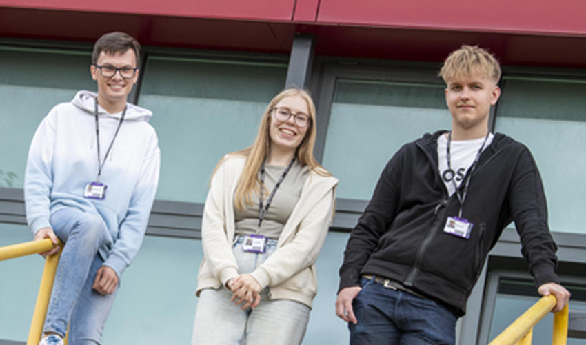 A female student in between two male students stood at the top of some stairs, leaning on a railing, looking and smiling at the camera.