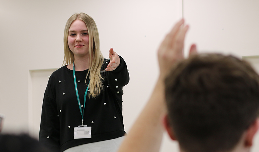 An image of a young student smiling and pointing to a member of the audience with their hand up. 