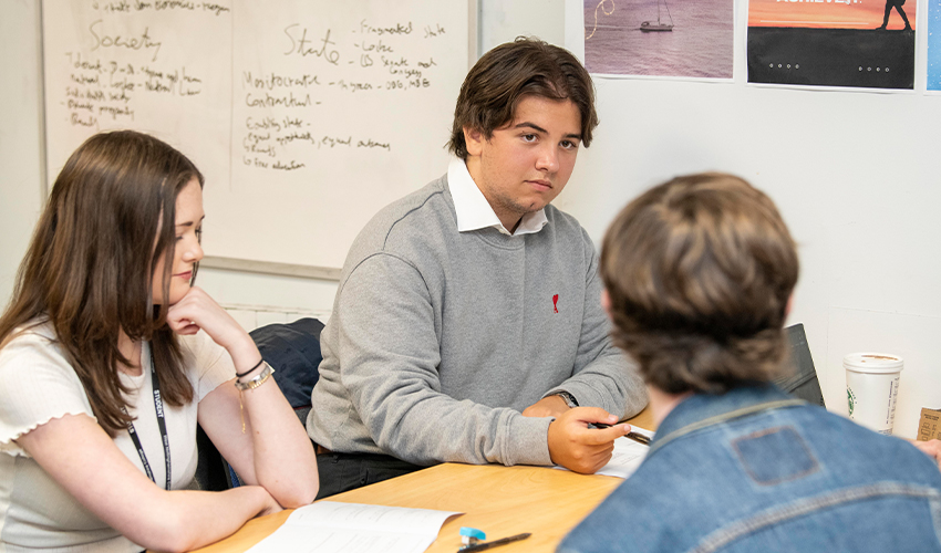 Three students in class together, sat at a desk, discussing classwork.