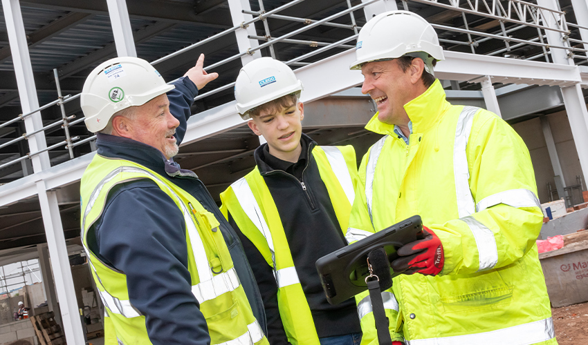 An image of a construction student looking at a laptop, in between two construction workers looking at each other and pointing to the building behind them